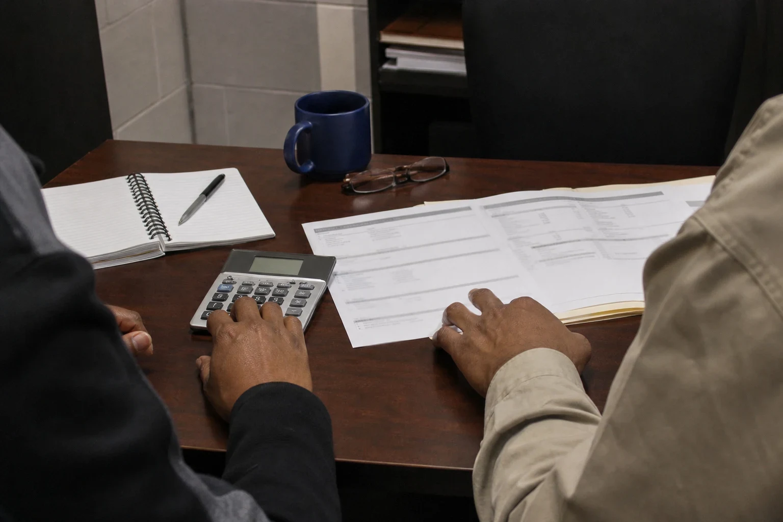 Two people at a wooden office desk, one entering figures on a calculator beside printed statements, with a spiral notepad, pen, blue mug and reading glasses